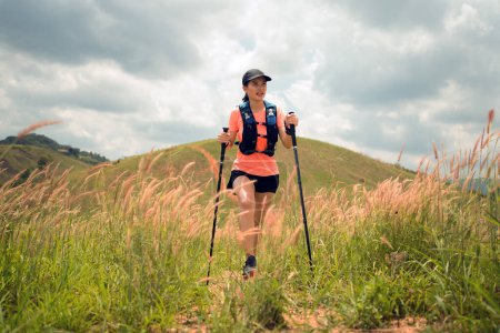 Téléchargez les photos : Sentier actif pour jeunes femmes traversant une prairie sur un sentier herbeux au sommet des montagnes l'après-midi avec perche de randonnée - en image libre de droit