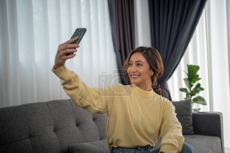 Photo for Asian woman sitting on sofa talking with friend via video call and selfie at home. - Royalty Free Image