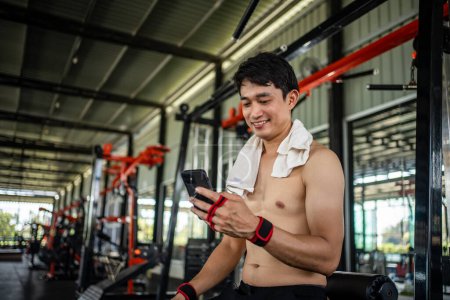 Photo for Young male playing phone and listening to music after exercise with various exercise equipment in fitness. - Royalty Free Image
