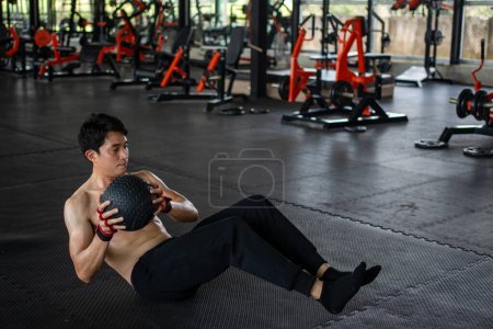 Téléchargez les photos : Muscular man exercising with a pilates ball at the gym. Male sitting on the floor and doing a workout using a medicine ball. - en image libre de droit