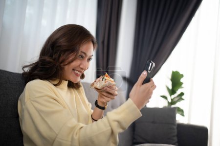 Foto de Mujer asiática usando videollamada telefónica con un amigo mientras come sándwich en casa - Imagen libre de derechos