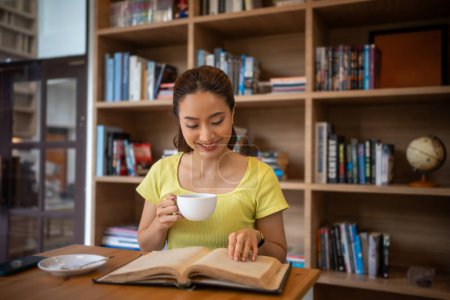 Photo for Young woman reading book and drink hot coffee in library at home, smiling, laughing, enjoying bookworms hobby, home leisure time - Royalty Free Image
