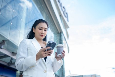 Foto de Joven empresaria sosteniendo una taza de café caliente y usando un teléfono inteligente mientras camina por la ciudad - Imagen libre de derechos