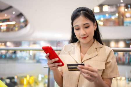 Photo for Young woman in smart casual wear carrying many paper shopping bags shopping online on mobile phone using credit card at shopping center - Royalty Free Image