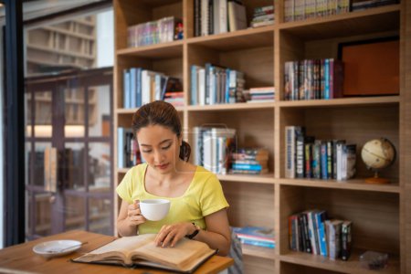 Téléchargez les photos : Jeune femme lisant un livre et buvant du café chaud à la bibliothèque à la maison, souriant, riant, appréciant les vers de bibliothèque passe-temps, loisirs à la maison - en image libre de droit