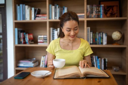 Foto de Mujer joven leyendo libro y beber café caliente en la biblioteca en casa, sonriendo, riendo, disfrutando de los gusanos de biblioteca hobby, tiempo libre en casa - Imagen libre de derechos