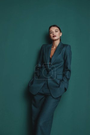 Photo for Fashionable confident woman wearing elegant green suit with blazer and trousers posing on green wall background. Studio fashion portrait - Royalty Free Image