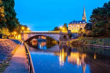 Téléchargez les photos : Les eaux tranquilles de la rivière Avon serpentent à travers la ville et le clocher de l'église St Johns s'élève dramatiquement contre un ciel bleu à Bath, Royaume-Uni - en image libre de droit