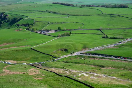 Luftaufnahme einer malerischen Landschaft mit saftig grünen Feldern, kurvenreichen Straßen und charmanten Landhäusern im Peak District Park, England.