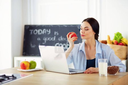 Téléchargez les photos : Femme avec pomme et ordinateur portable assis dans la cuisine
 . - en image libre de droit