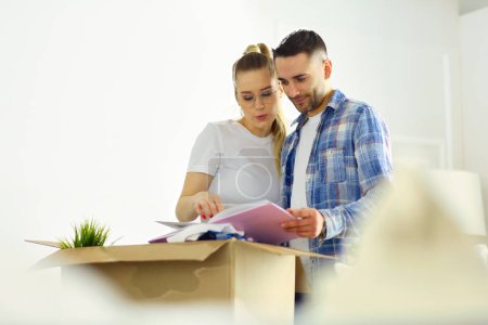 Téléchargez les photos : Un heureux mari et femme caucasiens d'âge moyen debout dans leur nouvelle maison et sortant l'album photo encadré de la boîte en carton, bavardant animément et riant - en image libre de droit
