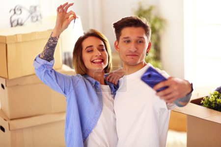 Photo for Young happy man and woman make selfie on smartphone, holding keys of new home after buying house apartment. - Royalty Free Image