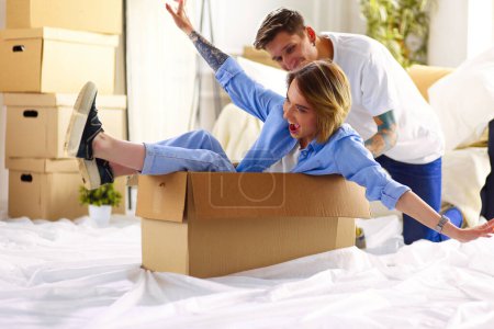 Photo for Couple having fun laughing moving into new home, young woman riding sitting in cardboard box while man pushing it. - Royalty Free Image