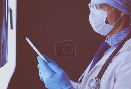 Photo for Doctor in hospital sitting at desk looking at x-rays on tablet against white background with x-rays. - Royalty Free Image