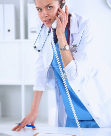 Photo for Portrait of young woman doctor in white coat at computer using phone - Royalty Free Image