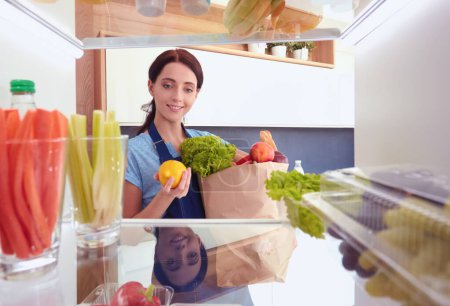 Foto de Retrato de una mujer parada cerca de una nevera abierta llena de alimentos saludables, verduras y frutas. Retrato de mujer. - Imagen libre de derechos