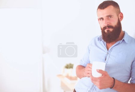 Téléchargez les photos : Beau jeune homme indien tenant une tasse de café et souriant tout en se tenant debout sur fond blanc
. - en image libre de droit