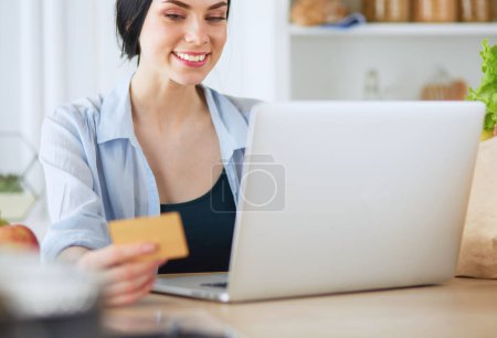 Foto de Mujer sonriente compras en línea utilizando la computadora y la tarjeta de crédito en la cocina
. - Imagen libre de derechos