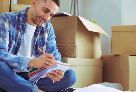 Photo for A moving man sitting on the floor in empty apartment, Among the Boxes, Checking the List of Things - Royalty Free Image