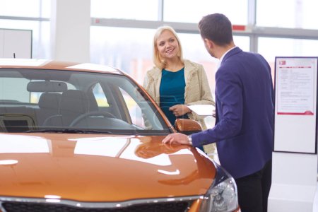 Photo for Dealer with woman stands near a new car in the showroom. - Royalty Free Image