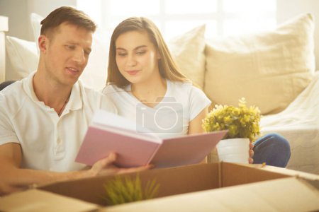 Photo for Young couple holding album while sitting on floor in new house. - Royalty Free Image