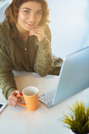 Téléchargez les photos : Belle femme souriante assise au bureau, tenant une tasse de café, elle se relaxe et regarde ailleurs. - en image libre de droit