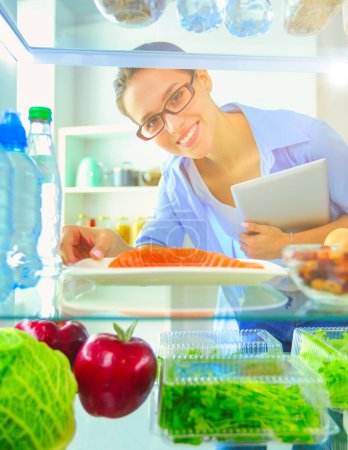 Téléchargez les photos : Portrait de femme debout près d'un réfrigérateur ouvert rempli d'aliments sains, de légumes et de fruits - en image libre de droit