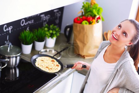 Photo for Young woman prepares pancakes in the kitchen while standing near the table. Woman in the kitchen. Cooking at kitchen - Royalty Free Image