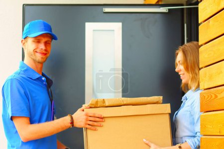 Foto de Repartidor sonriente con uniforme azul que entrega la caja de paquetes al destinatario: concepto de servicio de mensajería. Repartidor sonriente en uniforme azul. - Imagen libre de derechos