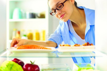 Téléchargez les photos : Portrait de femme debout près d'un réfrigérateur ouvert rempli d'aliments sains, de légumes et de fruits. - en image libre de droit