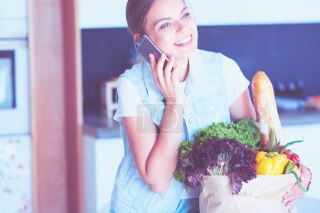 Foto de Mujer sonriente con teléfono móvil sosteniendo bolsa de compras en la cocina. - Imagen libre de derechos