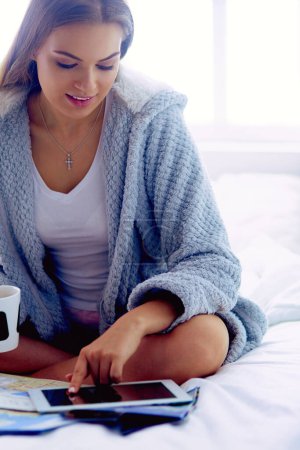 Photo for Relaxed young woman sitting on bed with a cup of coffee and digital tablet. - Royalty Free Image