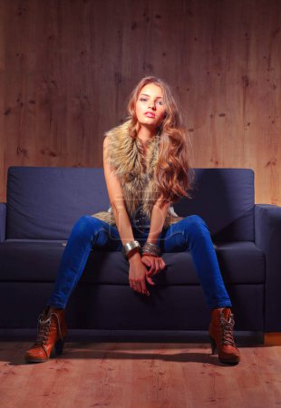 Téléchargez les photos : Portrait de femme élégante assise sur un canapé noir portant un jean bleu et un gilet de fourrure
. - en image libre de droit
