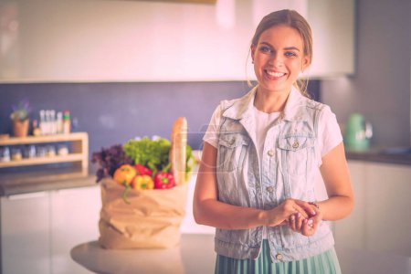 Téléchargez les photos : Jeune femme tenant sac d'épicerie avec des légumes. Debout dans la cuisine. - en image libre de droit