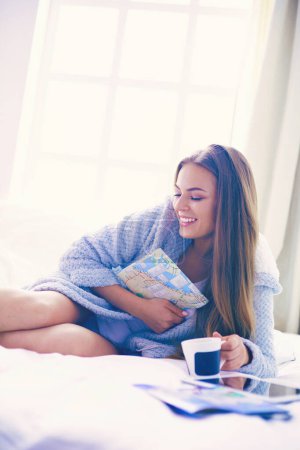 Téléchargez les photos : Jeune femme détendue assise sur le lit avec une tasse de café et une tablette numérique. - en image libre de droit