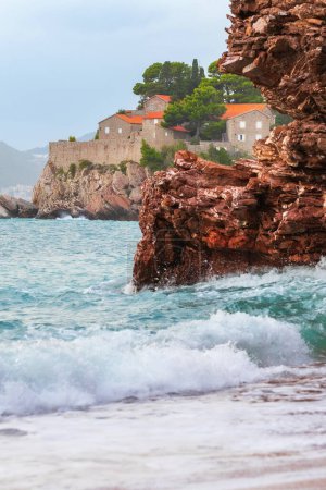 Foto de Hotel en la isla de Sveti Stefan al atardecer, cerca de Budva, Montenegro. Olas y rocas rojas - Imagen libre de derechos