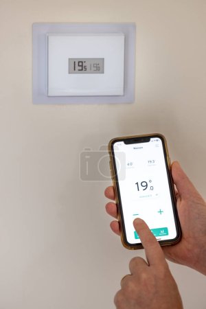 Photo for Person holding a smartphone in their hands to adjust their connected thermostat in a French house - Royalty Free Image