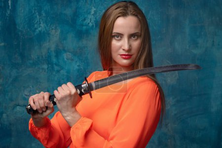 Foto de Modern female warrior. Beautiful woman in an orange dress with a samurai sword on a blue background. - Imagen libre de derechos