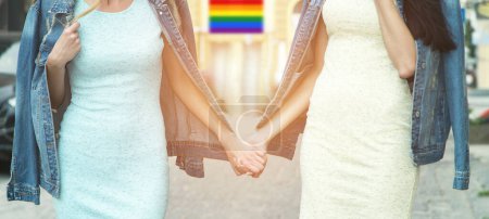 Two women hold hands and walk down the street, against the backdrop of a building with a flag of lgbt pride. Close-up of hands, faces are not visible.