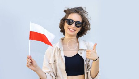 Hermosa mujer en gafas de sol sostiene la bandera de Polonia en sus manos y muestra un pulgar hacia arriba.