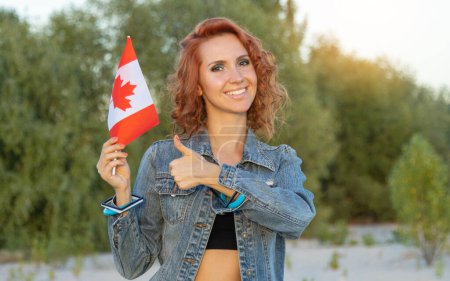 Foto de Mujer joven sonriente con bandera de Canadá mostrando los pulgares hacia arriba mientras está de pie en el fondo de la naturaleza. - Imagen libre de derechos