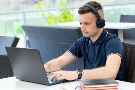 Male freelancer works at a laptop and listens to music. Student man watches a webinar and listens to headphones while sitting at a table in a co working cafe.