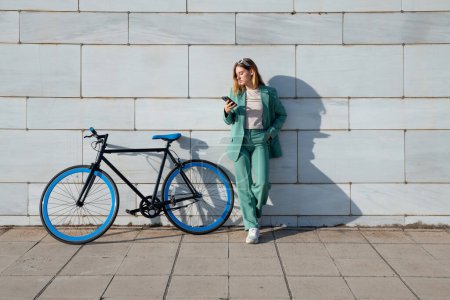 Téléchargez les photos : Jeune femme aux cheveux roux habilitée en costume vert en utilisant un smartphone debout avec son vélo sur fond de marbre - en image libre de droit