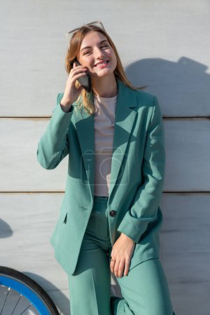 Téléchargez les photos : Jeune femme aux cheveux roux habilitée en costume vert parlant sur smartphone debout sur fond de marbre - en image libre de droit