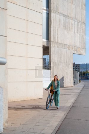 Téléchargez les photos : Jeune femme aux cheveux roux habilitée en costume vert se promenant à vélo dans le quartier financier avec un bâtiment moderne en arrière-plan - en image libre de droit