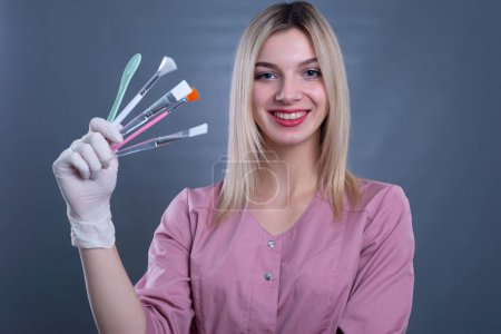 a young girl doctor holds cosmetic brushes for skin procedures in her hands. On a gray background.