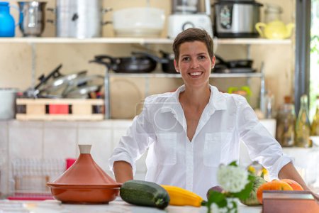 Relaxed woman with a pile of organic vegetables in front and blurry kitchen utensils in the back.