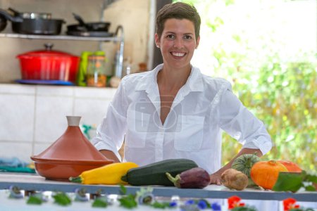 Junge lächelnde Frau hinter einer Arbeitsplatte, die Gemüse aus dem Gemüsegarten für die Tagine trägt.