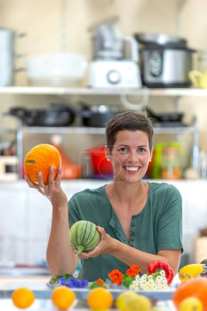 Mujer sonriente y relajada, con una pila de verduras orgánicas en frente y sosteniendo un calabacín y calabaza.