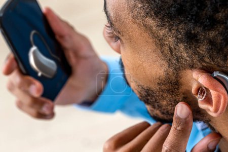 Téléchargez les photos : Jeune homme ajustant l'intensité du volume de l'aide auditive avec le smartphone. - en image libre de droit